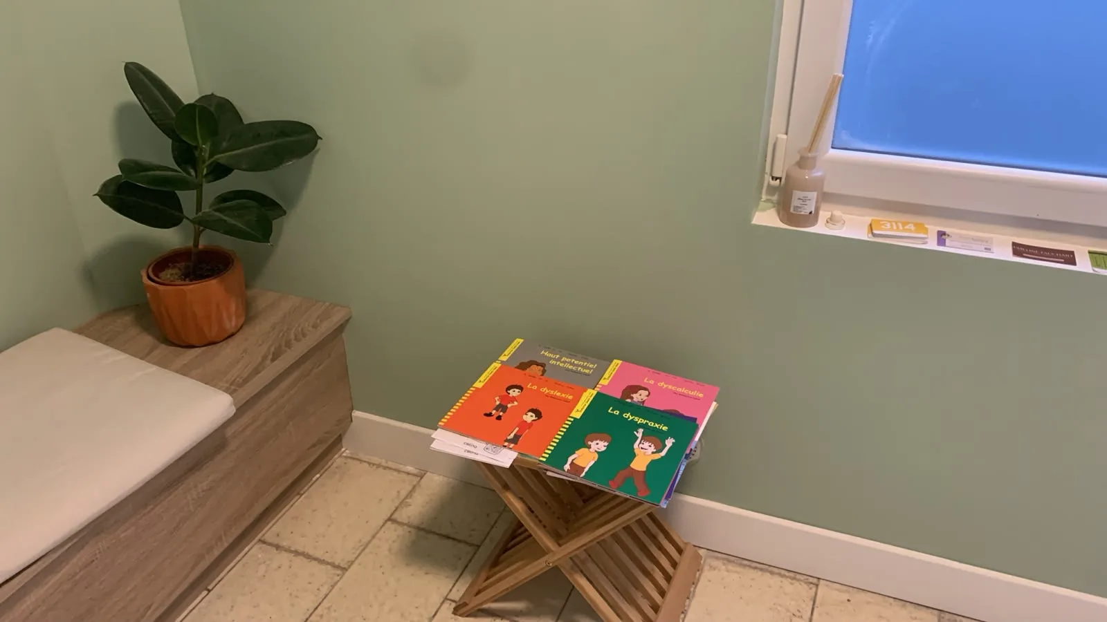 Coin salle d'attente avec une plante en pot terracotta, une table basse en bois pliable couverte de livres et une fenêtre lumineuse.