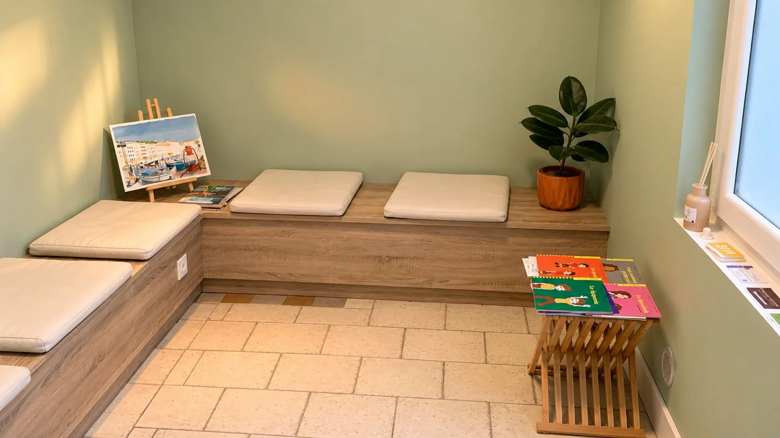 Vue d'ensemble de la salle d'attente : assises en bois avec coussins blancs, chevalet, table basse avec livres et grande fenêtre.